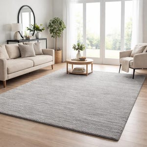 May include: A light gray rectangular area rug in a living room setting. The rug has a textured surface and is placed on a hardwood floor. The room also features a beige sofa, armchair, and a round coffee table.