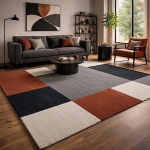 Puede incluir: Una alfombra rectangular grande con un patrón geométrico de bloques de colores sólidos en tonos naranja óxido, gris carbón, gris claro y crema. La alfombra tiene una superficie de pelo texturizado y está colocada sobre un suelo de madera.