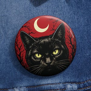 May include: A circular pinback button featuring a black cat with yellow eyes centered against a deep red background. The design includes a pale yellow crescent moon at the top and dark, silhouetted tree branches extending from the edges of the circle.