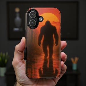 May include: A phone case featuring a Bigfoot silhouette in a sunset scene. The design shows the creature standing in water with its reflection, against a backdrop of an orange sun and trees. The case has a glossy finish.