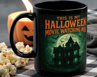 May include: Black ceramic Halloween mug with the text "THIS IS MY HALLOWEEN MOVIE WATCHING MUG" and a spooky haunted house design. A lit jack-o'-lantern candle and popcorn are in the background.
