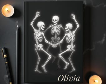 May include: Black journal featuring a dancing skeleton design. The illustration shows three skeletons holding hands, with the name "Olivia" in a cursive font. Two lit candles and a pen are on the dark surface.