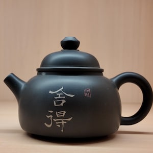 Könnte beinhalten: Schwarze Keramik-Teekanne mit rundem Körper, gebogenem Griff und Ausguss. Der Deckel hat einen kleinen Knopf. Chinesische Schriftzeichen sind auf der Vorderseite eingraviert. Die Teekanne wird wahrscheinlich zum Brühen von Tee verwendet.