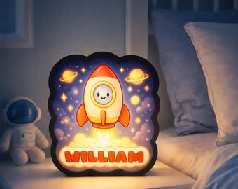 May include: A night light with a cartoon rocket ship design. The rocket is orange and white, with a smiling face. The name "WILLIAM" is displayed in orange letters. The background features planets and stars.