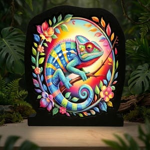 May include: A colorful chameleon illustration on a black, stone-shaped base. The chameleon is perched on a branch, surrounded by flowers and leaves. The artwork features vibrant blues, yellows, greens, and reds, creating a tropical theme.