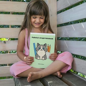 Puede incluir: Una persona sentada sostiene un cuaderno verde claro titulado "Palletta's Graph Notebook". La portada presenta un gato de dibujos animados, una regla y lápices. La persona lleva un atuendo rosa y está sentada en un banco de madera.