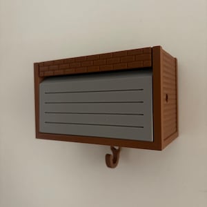 Może przedstawiać: Brązowy i szary miniaturowy haczyk ścienny z motywem garażu. Na górze znajduje się wzór cegieł, a drzwi garażowe są szare z poziomymi liniami. Mały haczyk wisi poniżej.