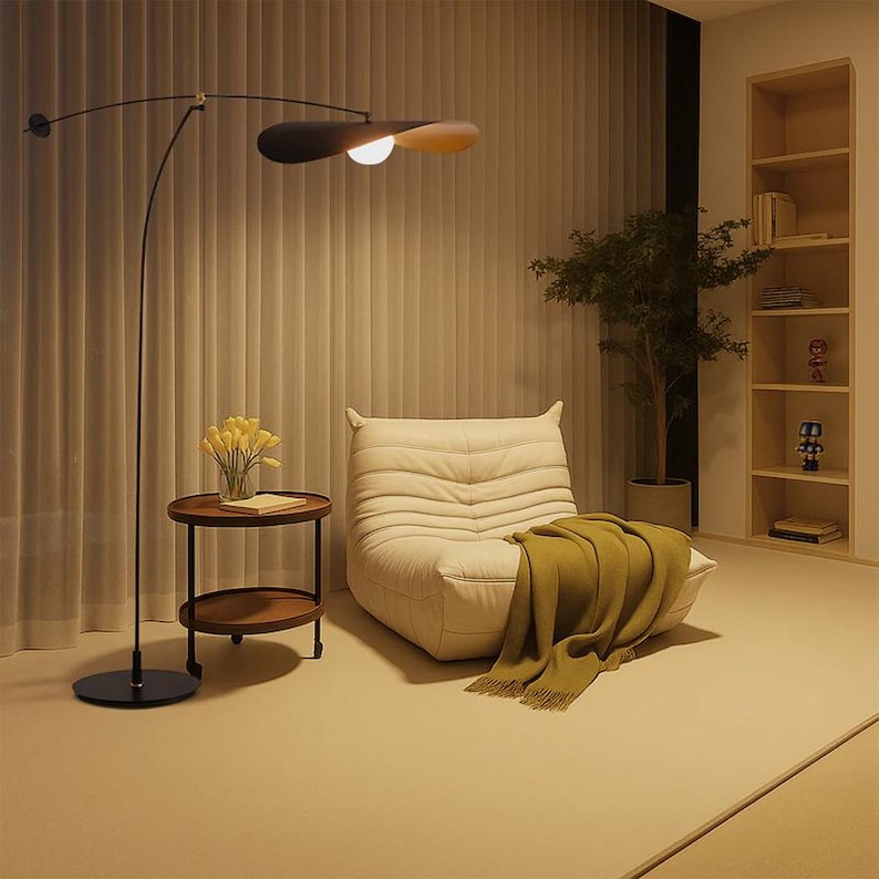 May include: A modern living room scene featuring a cream-colored armchair, a round side table with yellow flowers, and a unique arc floor lamp with a black and wood-toned shade. A green blanket is draped over the chair.