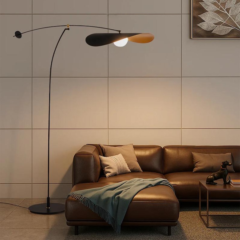 May include: A modern black floor lamp with a curved arm and a unique shade design. The shade is a combination of black and tan, with a round light bulb. The lamp is positioned next to a brown leather sofa.