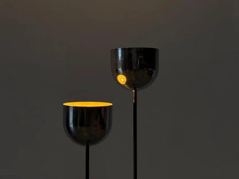 May include: Two floor lamps with tall, thin black stems and rounded, bowl-shaped heads. The exterior of the lamp heads is a reflective, dark metallic finish, while the interior glows with a warm, bright yellow light, creating a contrast.