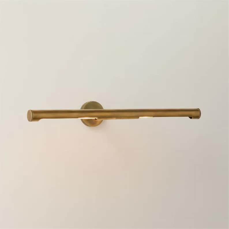 May include: A brass-toned picture light fixture. The light has a horizontal bar design with a circular base. The fixture is mounted on a white wall. The light is on, illuminating the area below.