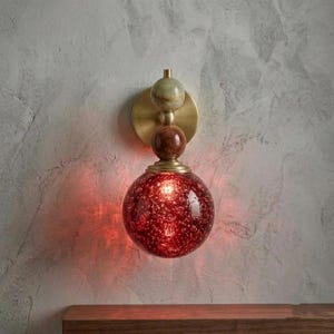 May include: A wall sconce featuring a circular brass backplate, a small green marble sphere, a medium brown wooden sphere, and a large red glass globe with a textured, bubbly interior finish that emits a warm, glowing light from within.