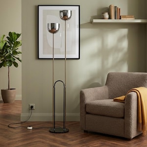 May include: A floor lamp featuring two chrome bowl-shaped shades mounted on slender vertical poles of varying heights. The lamp has a black circular base and a black arched support structure, connected to a black power cord with a floor switch.