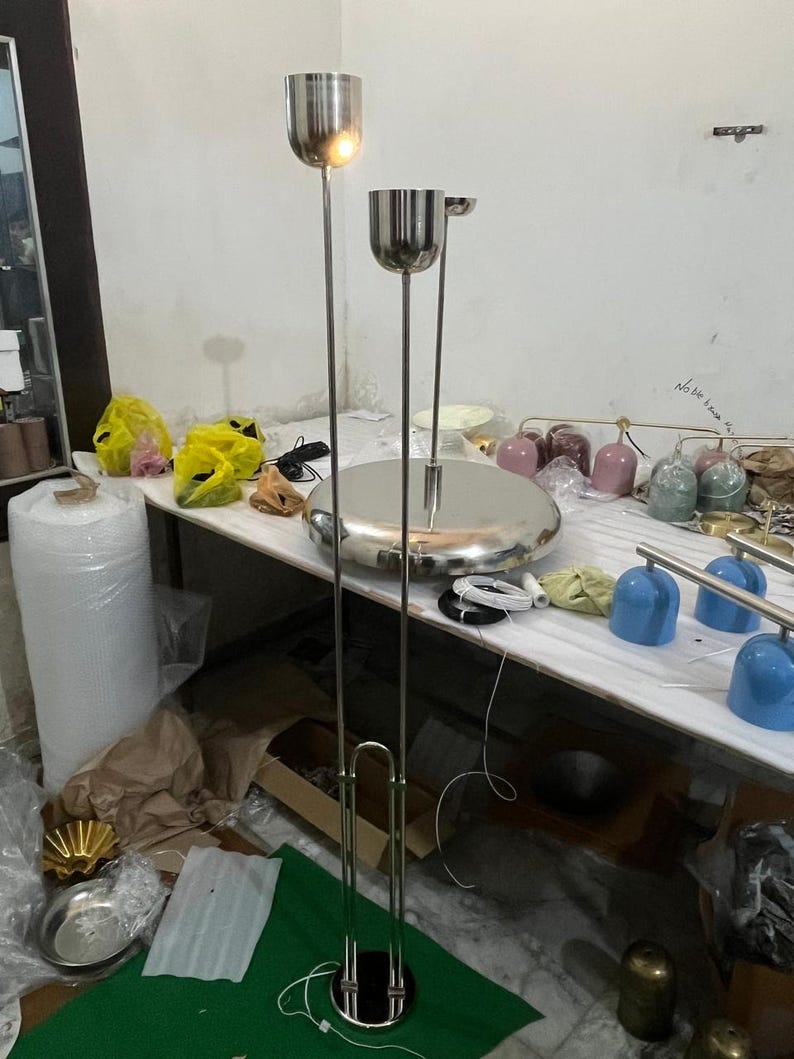 May include: A tall floor lamp featuring a polished silver metal finish with two vertical poles extending from a circular base. The lamp has two cylindrical light fixtures at different heights, each with a smooth, reflective surface and a clean design.