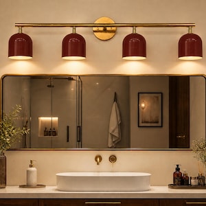 Può includere: Un moderno allestimento per il bagno con uno specchio rettangolare con cornice dorata, un apparecchio a quattro luci con tonalità bordeaux e un lavabo ovale bianco. Il mobile include un mobile in legno e oggetti decorativi.