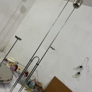 May include: A tall, silver-toned floor lamp featuring a circular base and two vertical metal poles of varying heights. Each pole is topped with a cylindrical, metallic shade. The structure is made of polished metal, showcasing a sleek, industrial design.
