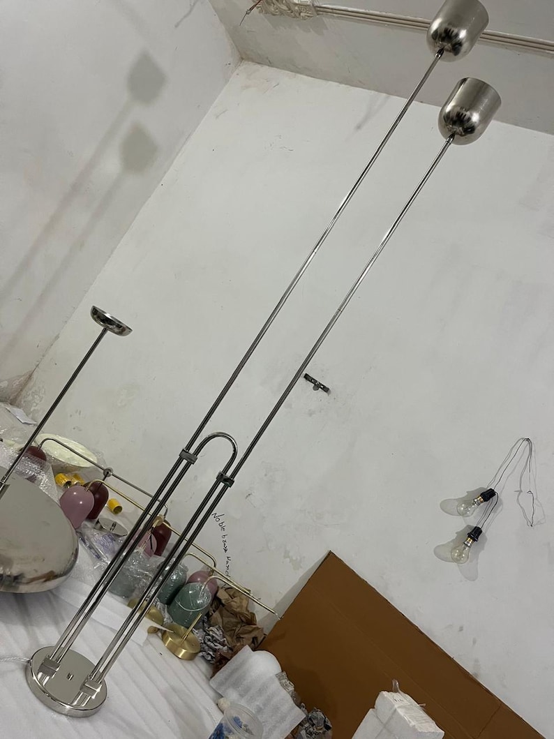 May include: A tall, silver-toned floor lamp featuring a circular base and two vertical metal poles of varying heights. Each pole is topped with a cylindrical, metallic shade. The structure is made of polished metal, showcasing a sleek, industrial design.