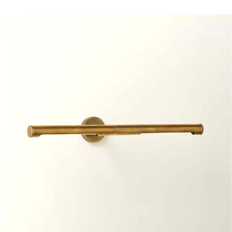 May include: A brass-colored towel bar mounted on a white wall. The bar is a horizontal cylinder with rounded ends, attached to a circular wall mount. The finish has a warm, metallic tone, suggesting a vintage or industrial aesthetic.