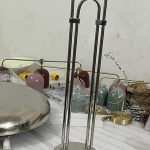 May include: A tall, silver-toned metal lamp stand with a circular base and two parallel vertical rods connected by a curved top piece. The text "Noble brass Hand" is written on the surface behind the lamp. Several colorful lamp shades are visible.