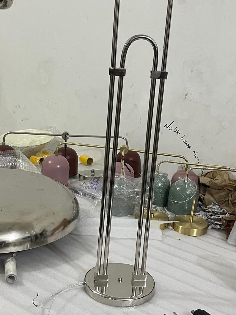 May include: A tall, silver-toned metal lamp stand with a circular base and two parallel vertical rods connected by a curved top piece. The text "Noble brass Hand" is written on the surface behind the lamp. Several colorful lamp shades are visible.