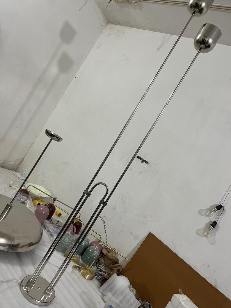 May include: A tall floor lamp featuring a polished silver-tone metal finish with two slender vertical poles connected by a U-shaped bracket. The lamp has two cylindrical light heads at the top and a circular base, providing a sleek and modern appearance.