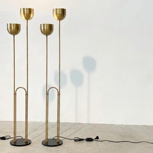 May include: Two tall floor lamps made of brushed brass, each featuring two slender vertical stems of varying heights topped with hemispherical bowl-shaped shades. The lamps are supported by circular black bases and include black power cords.
