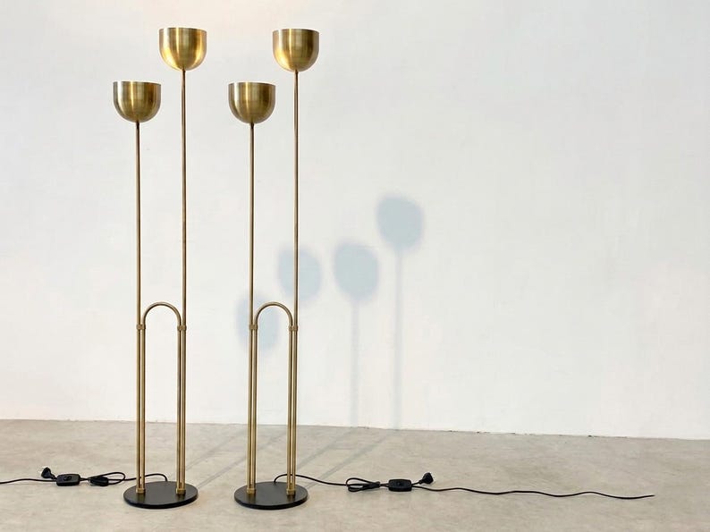 May include: Two tall floor lamps made of brushed brass, each featuring two slender vertical stems of varying heights topped with hemispherical bowl-shaped shades. The lamps are supported by circular black bases and include black power cords.