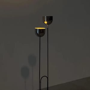 May include: A floor lamp featuring two thin, vertical black metal poles of different heights, each topped with a polished chrome bowl-shaped shade. The interior of the lower shade emits a warm, glowing yellow light, while the upper shade is dark.