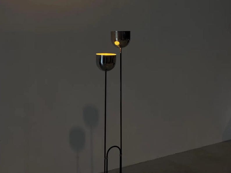 May include: A floor lamp featuring two thin, vertical black metal poles of different heights, each topped with a polished chrome bowl-shaped shade. The interior of the lower shade emits a warm, glowing yellow light, while the upper shade is dark.