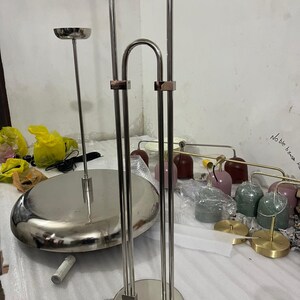 May include: A silver-toned metal floor lamp structure featuring a circular base and two vertical poles connected by a curved top piece. Beside it sits a large, flat, circular silver-toned metal lamp base and several small, colorful dome-shaped lamp shades.