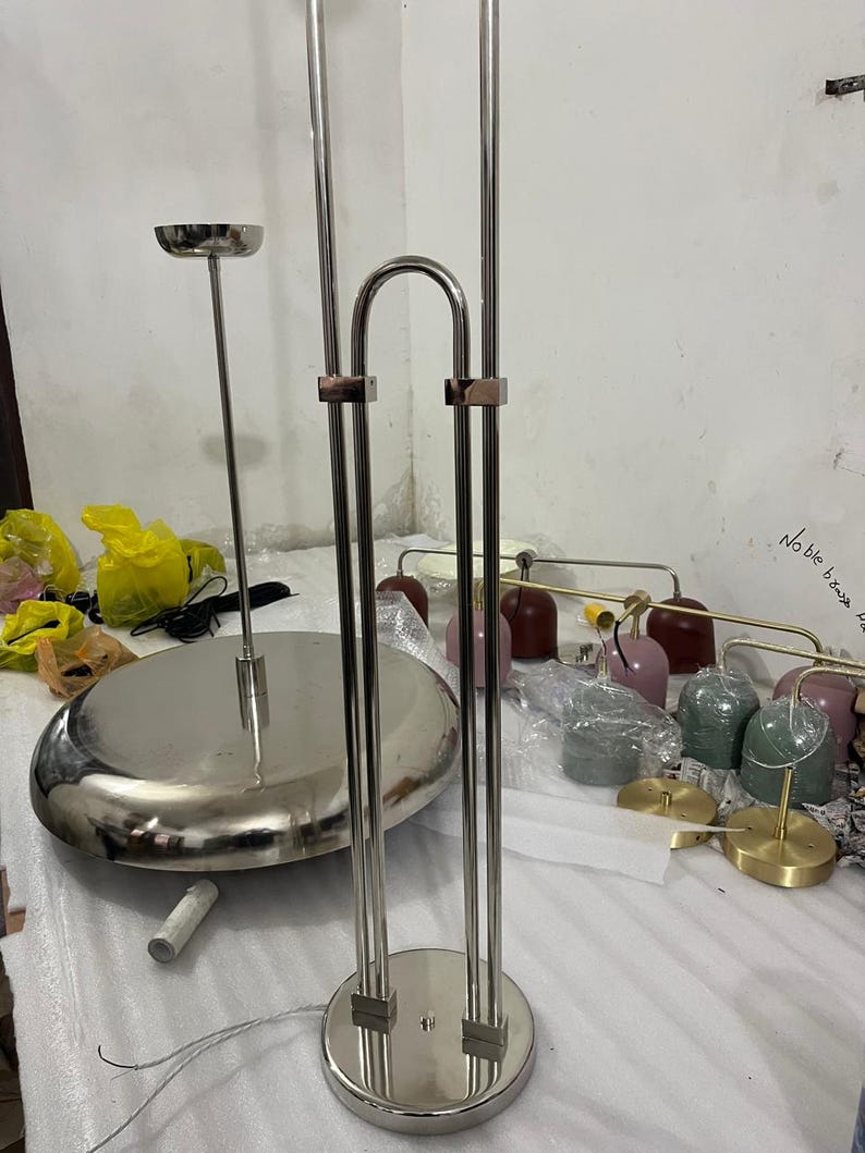 May include: A silver-toned metal floor lamp structure featuring a circular base and two vertical poles connected by a curved top piece. Beside it sits a large, flat, circular silver-toned metal lamp base and several small, colorful dome-shaped lamp shades.