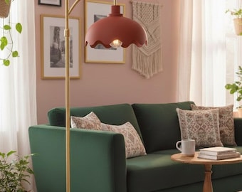 Puede incluir: Una lámpara de pie con base y mástil dorados, rematada con una pantalla festoneada de color óxido. La lámpara ilumina un salón con un sofá verde, cojines estampados y obras de arte enmarcadas.