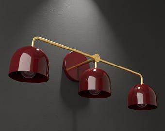 May include: A modern wall-mounted light fixture with three burgundy-colored, bell-shaped shades. The shades are attached to a gold-colored arm and a circular wall mount. The fixture is designed for indoor use.