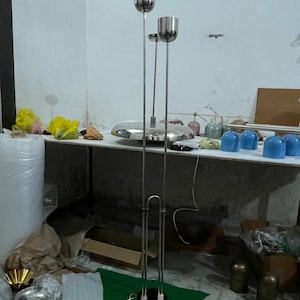 May include: A tall, silver-colored metal floor lamp featuring three slender vertical poles of varying heights, each topped with a cylindrical light fixture. The base is circular and connects to the poles, which are supported by a U-shaped metal frame.