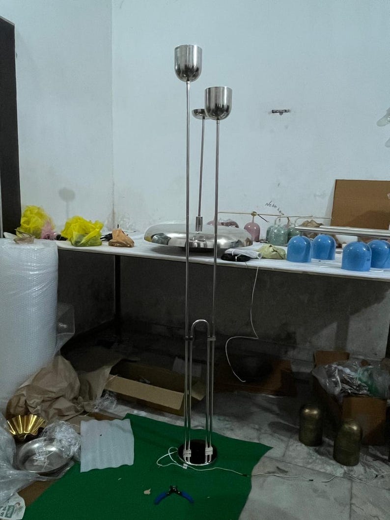May include: A tall, silver-colored metal floor lamp featuring three slender vertical poles of varying heights, each topped with a cylindrical light fixture. The base is circular and connects to the poles, which are supported by a U-shaped metal frame.
