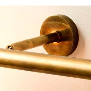 May include: A close-up of a brass-colored wall-mounted towel bar. The bar has a circular mounting plate with a ridged edge, a short arm, and a long, cylindrical bar. The finish is a warm, antique brass.