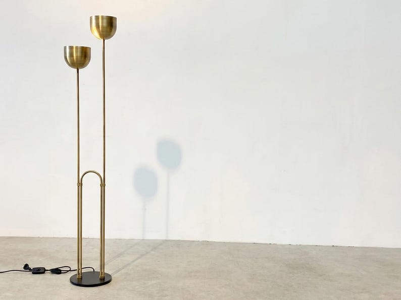 May include: A floor lamp featuring a circular black base and two vertical brass poles of varying heights. Each pole is topped with a hemispherical brass shade. The poles are connected by a central arched brass support structure near the base.