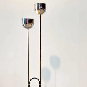 May include: A floor lamp featuring two polished chrome bowl-shaped shades mounted on slender vertical metal stems of unequal heights. The stems are connected at the base by a black U-shaped metal frame, creating a tall, dual-light structure.