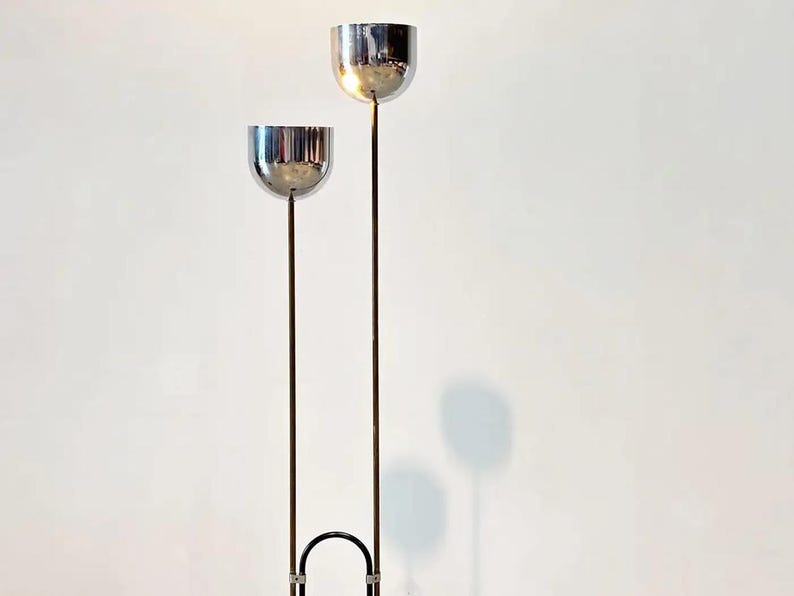May include: A floor lamp featuring two polished chrome bowl-shaped shades mounted on slender vertical metal stems of unequal heights. The stems are connected at the base by a black U-shaped metal frame, creating a tall, dual-light structure.