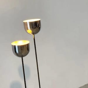 May include: Two floor lamps with polished chrome, bowl-shaped shades mounted on thin, dark metal stems. The lamps emit a warm, glowing light from within the rounded, reflective heads, which are positioned at different heights against a plain background.