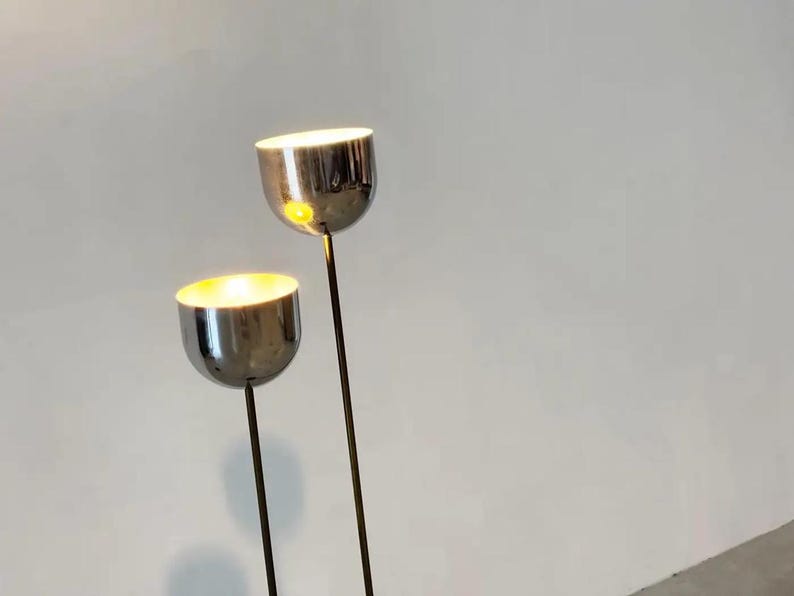 May include: Two floor lamps with polished chrome, bowl-shaped shades mounted on thin, dark metal stems. The lamps emit a warm, glowing light from within the rounded, reflective heads, which are positioned at different heights against a plain background.