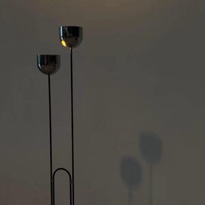 May include: A floor lamp featuring two thin, vertical black metal poles of unequal height, each topped with a polished chrome spherical shade. The poles are connected at the base by a curved metal arch, creating a tall, slender silhouette.