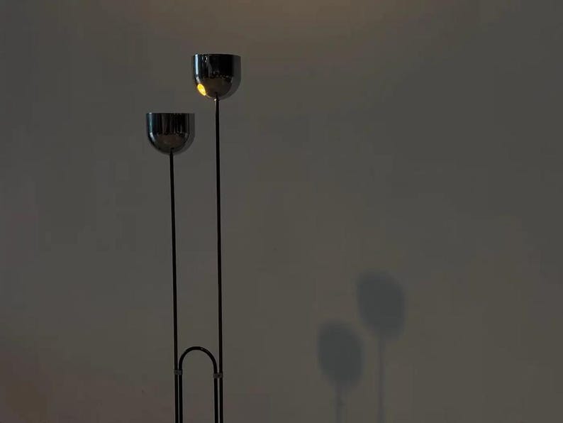 May include: A floor lamp featuring two thin, vertical black metal poles of unequal height, each topped with a polished chrome spherical shade. The poles are connected at the base by a curved metal arch, creating a tall, slender silhouette.