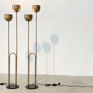 May include: Two tall floor lamps made of brushed brass metal, each featuring two slender vertical stems of varying heights topped with hemispherical bowl-shaped shades. The lamps stand on circular black bases and include black power cords.
