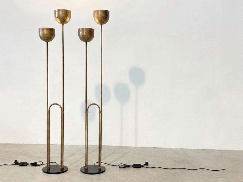 May include: Two tall floor lamps made of brushed brass metal, each featuring two slender vertical stems of varying heights topped with hemispherical bowl-shaped shades. The lamps stand on circular black bases and include black power cords.