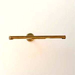 May include: A brass-toned, horizontal wall-mounted light fixture. The fixture has a cylindrical bar with a circular mounting plate. The light is positioned to shine downward, creating a warm glow. The design is minimalist and modern.