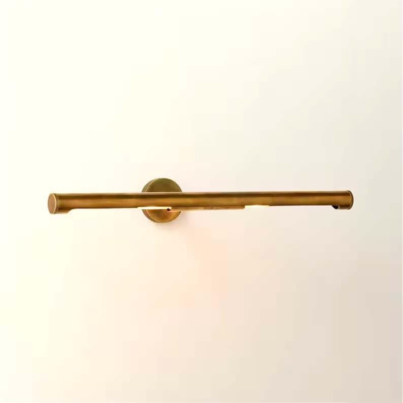 May include: A brass-toned, horizontal wall-mounted light fixture. The fixture has a cylindrical bar with a circular mounting plate. The light is positioned to shine downward, creating a warm glow. The design is minimalist and modern.