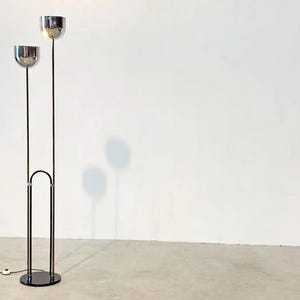 May include: A floor lamp featuring two chrome-finished bowl-shaped shades mounted on thin vertical poles of varying heights. The poles are connected by a black arched frame that extends from a circular black base, with a black power cord attached.
