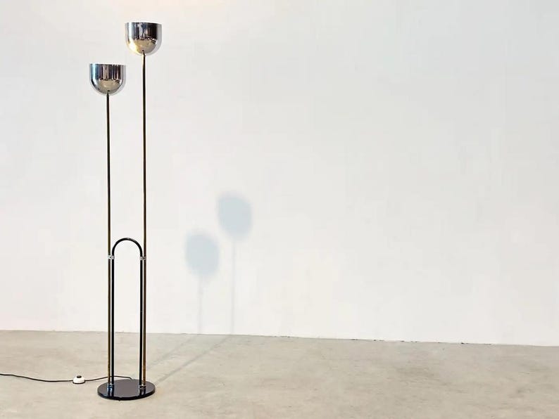 May include: A floor lamp featuring two chrome-finished bowl-shaped shades mounted on thin vertical poles of varying heights. The poles are connected by a black arched frame that extends from a circular black base, with a black power cord attached.