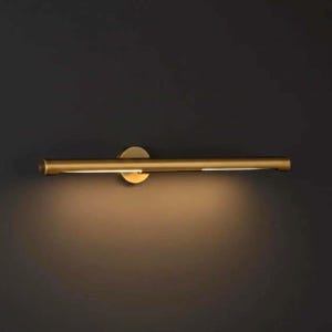 May include: A gold-colored, horizontal wall-mounted light fixture. The fixture has a cylindrical bar with a thin, bright light strip. It is attached to a circular base, casting a warm glow on the wall below. The background is a dark gray.